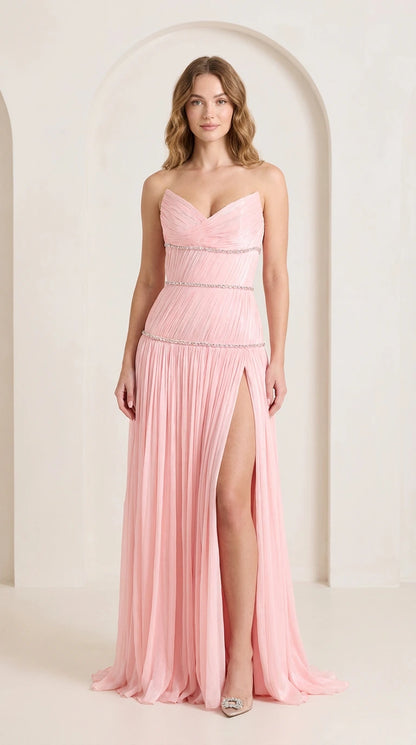 Front view of woman wearing strapless blush pink pleated evening gown with rhinestone trim and high slit