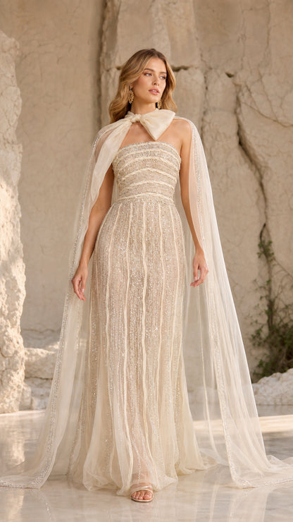 Front view of champagne strapless beaded maxi gown with flowing sheer cape and bow collar