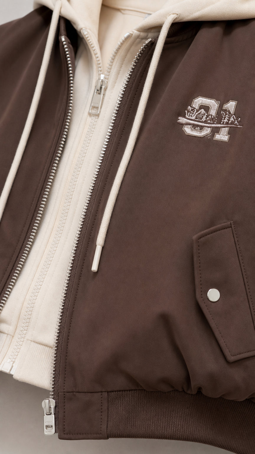 Close-up detail of brown bomber jacket showing double zipper, hood drawstring, and chest embroidery